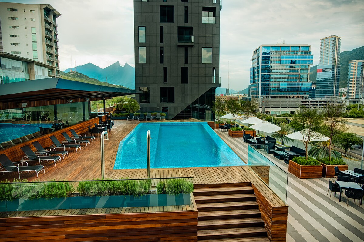 A modern building overlooks a tranquil swimming pool surrounded by wooden decking and lounge chairs. Lush greenery is visible along the edges, while nearby urban structures create a lively backdrop against the horizon. The sky features soft clouds, enhancing the serene atmosphere.