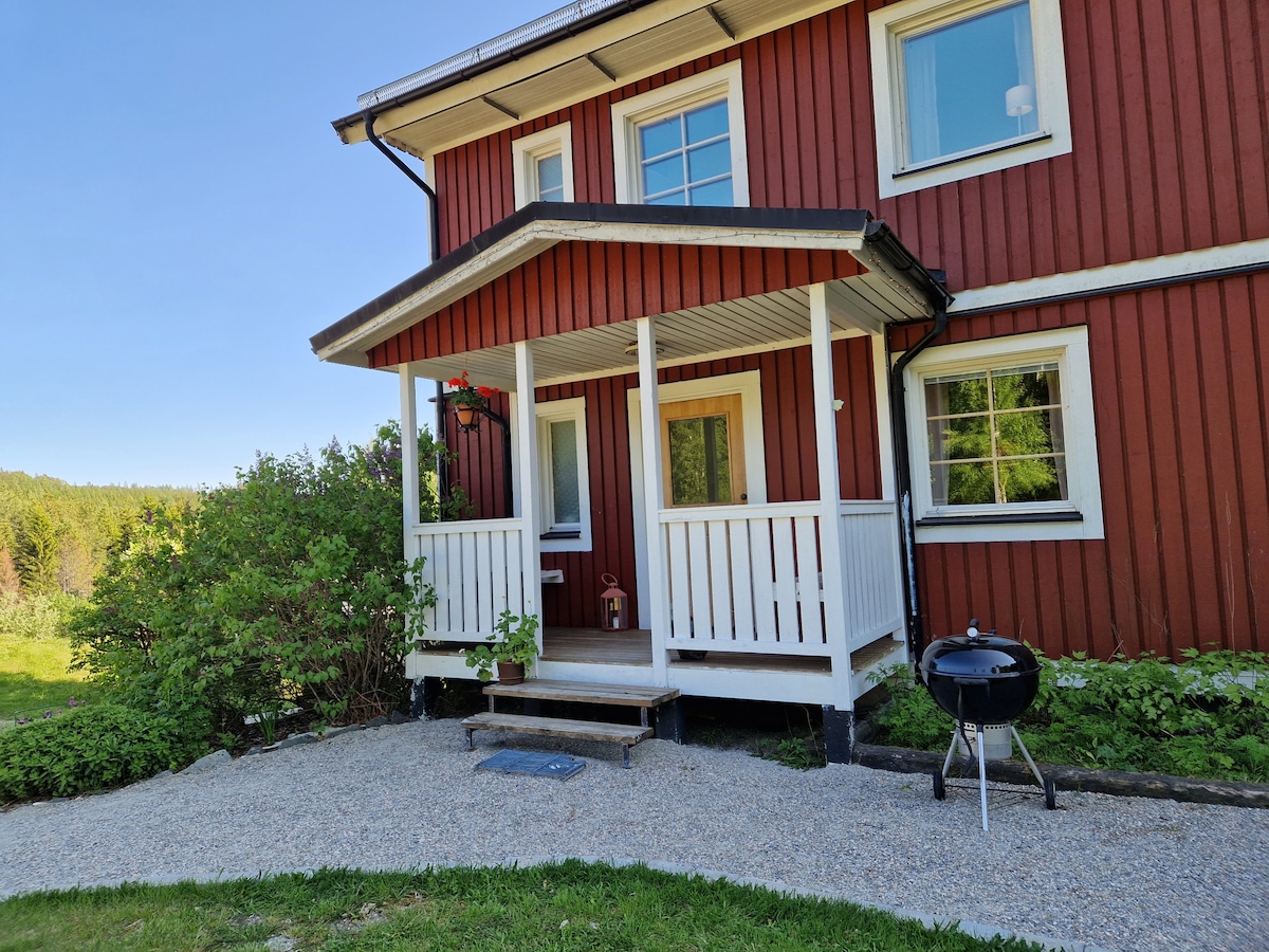 A red wooden house features a welcoming porch with white railings. A small charcoal grill is positioned nearby on the gravel pathway, surrounded by greenery. Large windows allow natural light while offering views of the outdoor landscape.