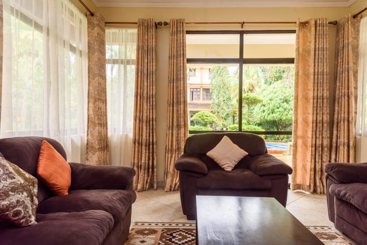 A spacious living area is highlighted by large windows allowing natural light to flood the room. Comfortable sofas, including one with an orange cushion, surround a wooden coffee table. Views of greenery and an outdoor pool are visible through the glass doors.