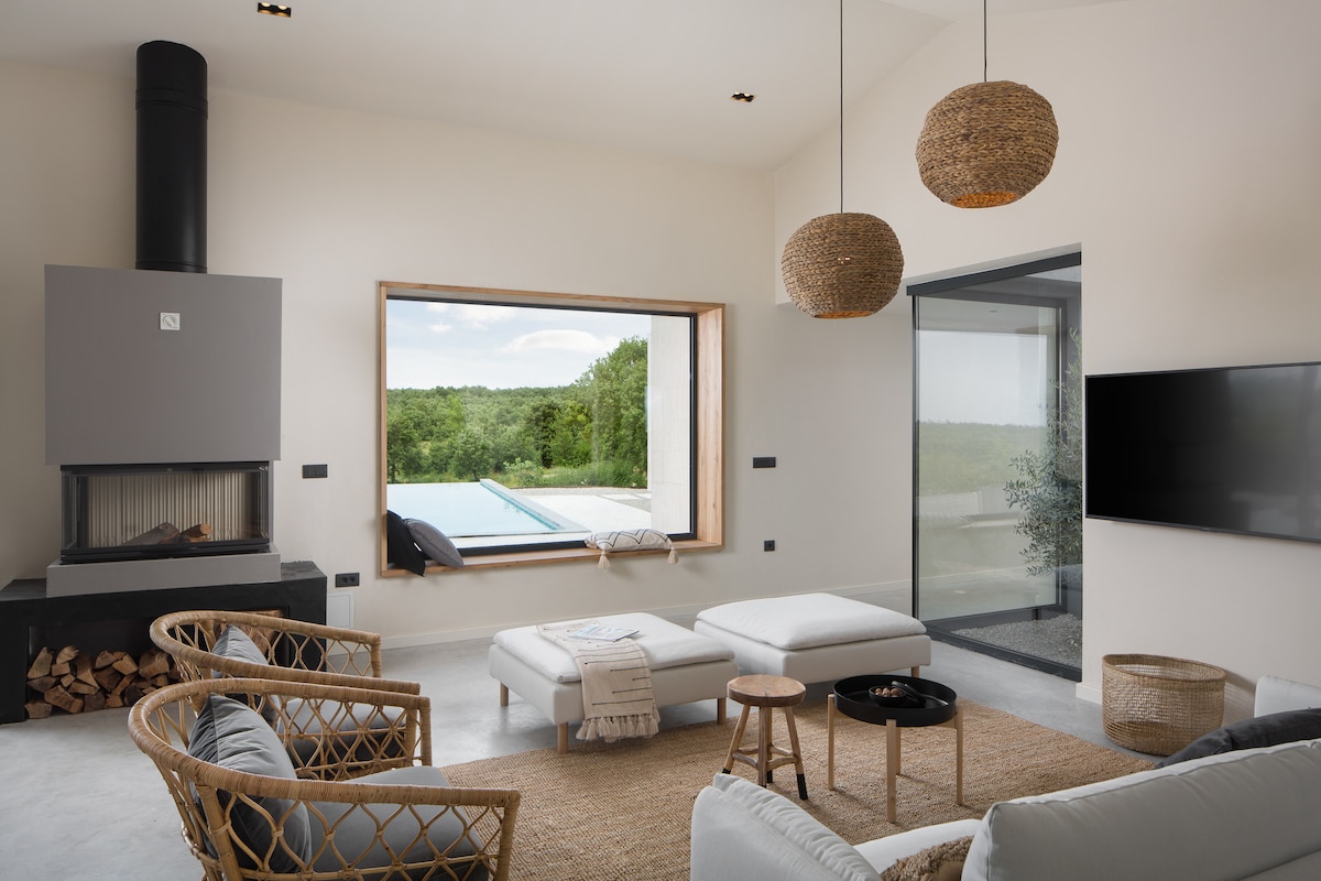 A welcoming living area features a large window that frames serene views of the surrounding greenery and the private pool. Cozy seating is arranged around a central table, and natural materials like rattan and neutral tones create a calming atmosphere.