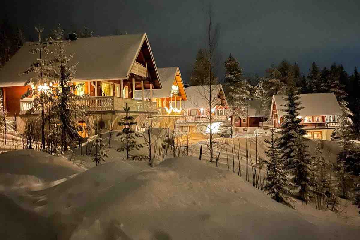 The exterior of several chalets is illuminated at night, surrounded by snow-covered trees. Soft lighting enhances the wood features of the buildings, showcasing their architectural style. The peaceful winter landscape is complemented by a cover of fresh snow, creating a serene atmosphere.