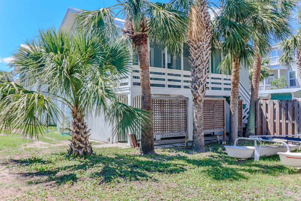 A lush outdoor area features vibrant palm trees swaying in a gentle breeze, providing a tropical feel. The cottage is partially visible above, showcasing a wraparound screened porch. A clean, grassy space is present, with outdoor storage tucked behind a lattice panel.