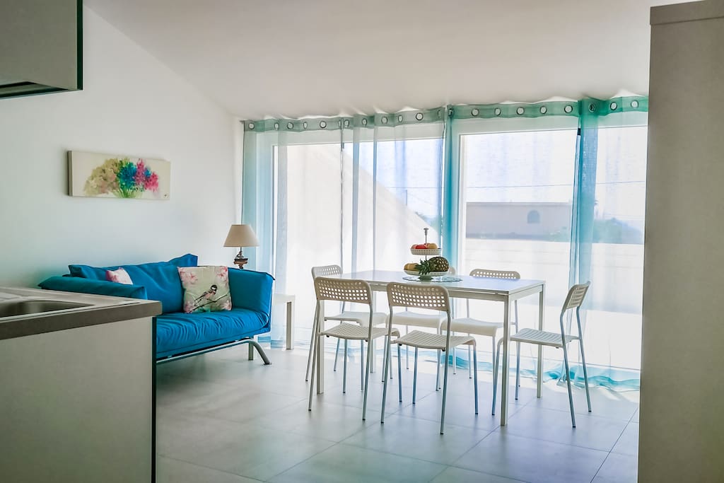 Cucina-soggiorno con ampie vetrate, accessibile alla veranda con vista mare/giardino, corredata di pergola e divanetti.