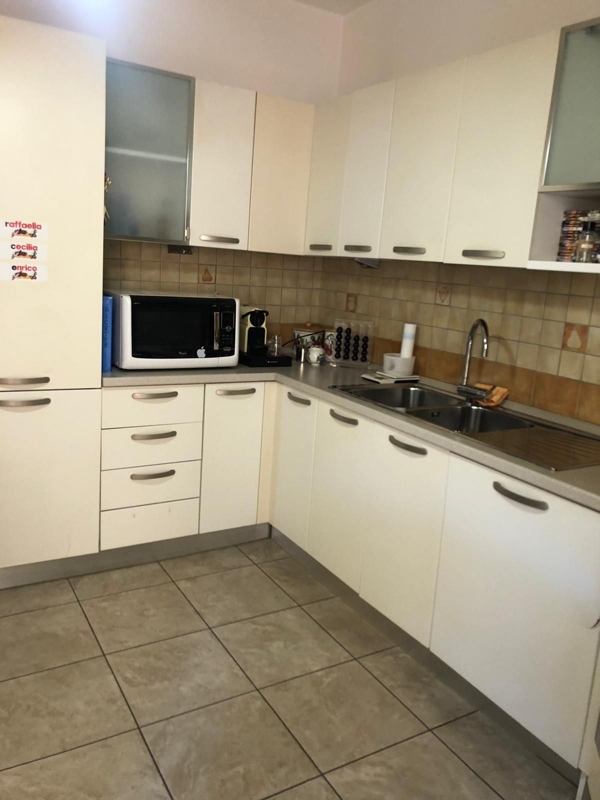 The kitchen features a modern design with light-colored cabinetry and tiled flooring. Essential appliances, including a microwave and coffee machine, are visible. Counter space is provided for meal preparation, and various utensils and condiments can be seen arranged on the shelves.