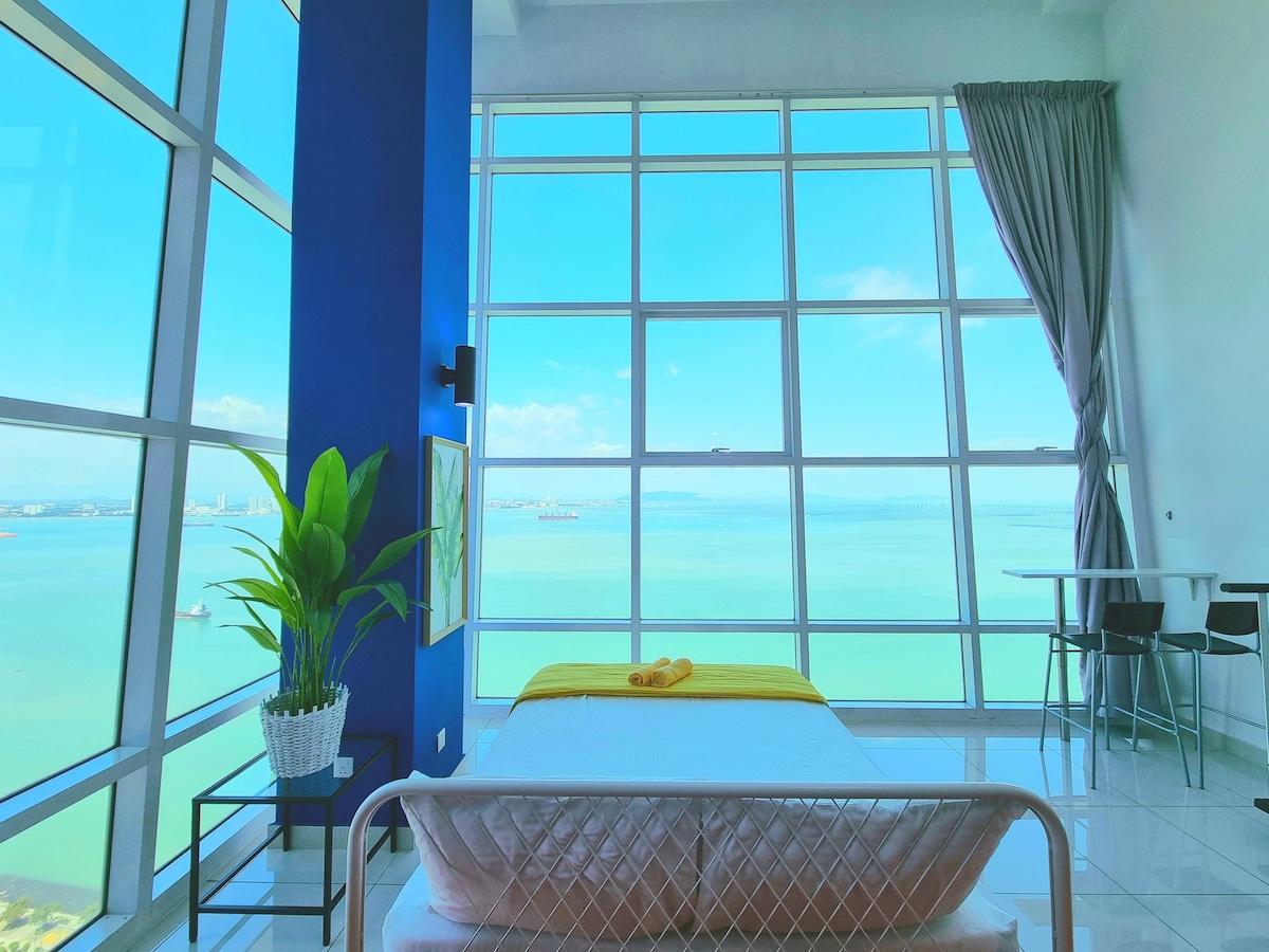 A spacious bedroom features a large window offering an unobstructed sea view. Natural light floods the room, while a comfortable bed with a neatly arranged yellow accent is positioned prominently. A potted plant adds a touch of greenery beside the window.