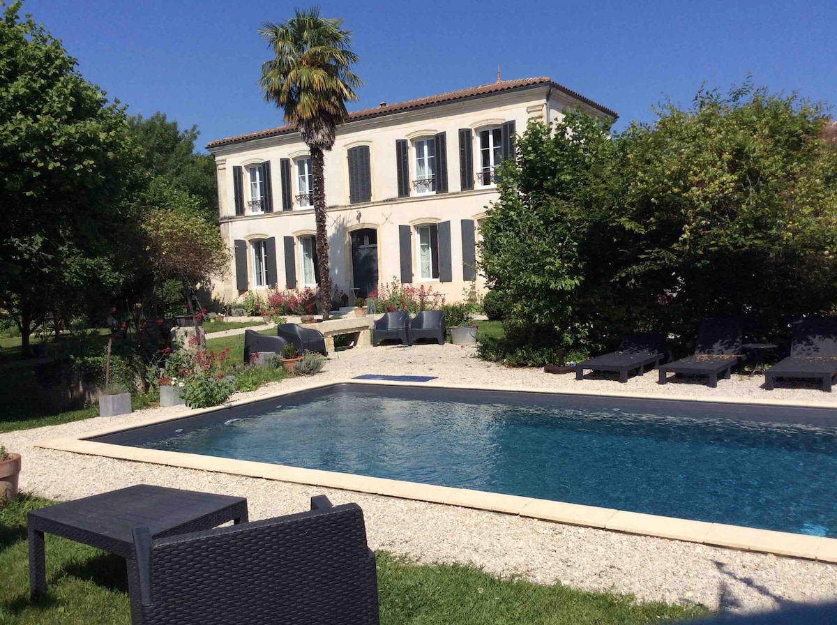 A spacious pool is set within a landscaped garden, surrounded by lush greenery and flowering plants. Lounge chairs are positioned alongside the water, offering relaxation opportunities. A charming two-story residence with dark-shuttered windows and a classic facade is visible in the background.