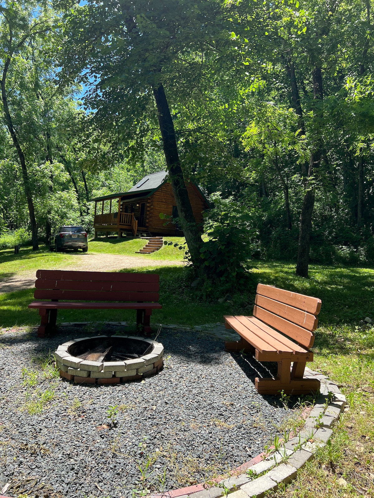 A tranquil outdoor area features two wooden benches positioned around a fire pit made of stone. Lush green trees provide shade, and a rustic cabin is visible in the background, nestled among the foliage. A gravel path leads to the cabin from the parking area.
