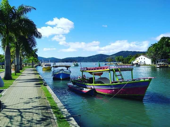 Colorful fishing boats are anchored along a calm waterway, surrounded by lush greenery and rolling hills. A paved pathway runs alongside, offering a scenic view of the serene environment, with bright blue sky and scattered clouds enhancing the tranquil atmosphere.