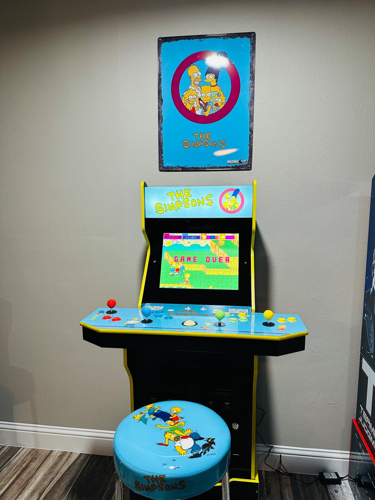 A colorful arcade game machine is displayed in a game room, featuring 'The Simpsons' theme. The screen shows a vibrant game scene, and the console includes various buttons and joysticks. A themed solid blue stool with a character design is positioned in front of the machine.