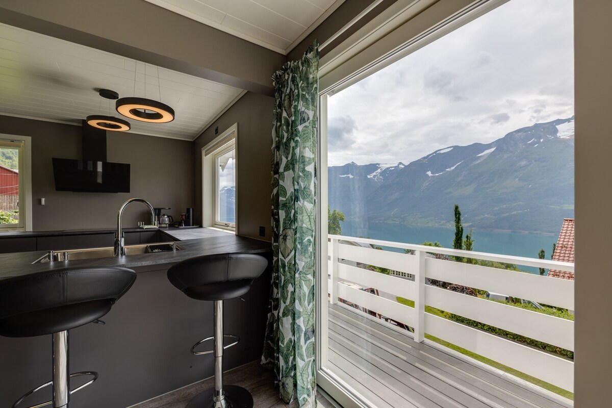 The kitchen area features a modern design with a sleek island and two bar stools. Large glass doors open to a scenic view of mountains and a lake, with natural light illuminating the space.