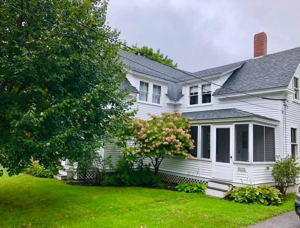 A charming two-story home is framed by a lush green lawn and several trees, with a flowering bush at the front. The white exterior features multiple windows and a screened porch. The grey roof complements the overall aesthetic, set against a cloudy sky.