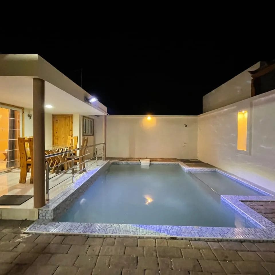 An illuminated pool area is surrounded by a sleek terrace, featuring a mosaic tile design. The space is complemented by modern outdoor furniture, and warm lights highlight the walls of a nearby dining area, creating a serene evening ambiance.