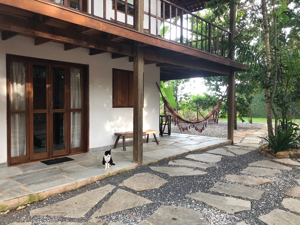 An outdoor area features a stone pathway leading to a wooden structure with a spacious balcony. A hammock hangs under the balcony, inviting relaxation. A black and white cat sits beside a bench near the entrance, surrounded by greenery and tropical trees.