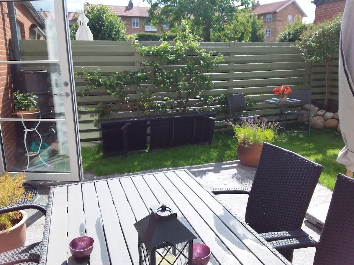 A private outdoor space is featured, showcasing a well-maintained garden surrounded by a wooden fence. A table with dark wicker chairs sits on a stone patio. Lush green grass, potted plants, and a decorative lantern are visible, enhancing the peaceful atmosphere.