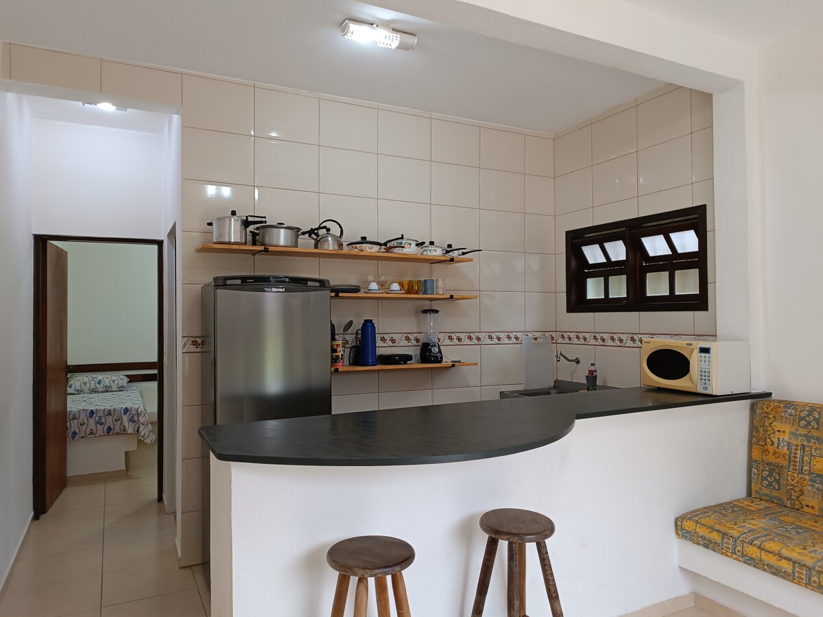 A clean and functional kitchen area is highlighted by a sleek countertop and wooden stools. Shelves display essential cooking utensils and appliances, while a large refrigerator stands against the wall. Natural light enters through a nearby window, creating a bright and inviting atmosphere.