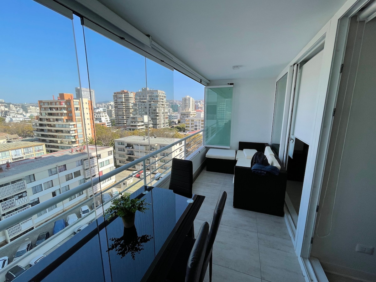 A modern balcony area offers a sleek dining table and comfortable seating, providing views of the cityscape. Large glass panels allow natural light to enter, creating an airy environment. The balcony is adorned with a small plant, contributing to the inviting appearance.
