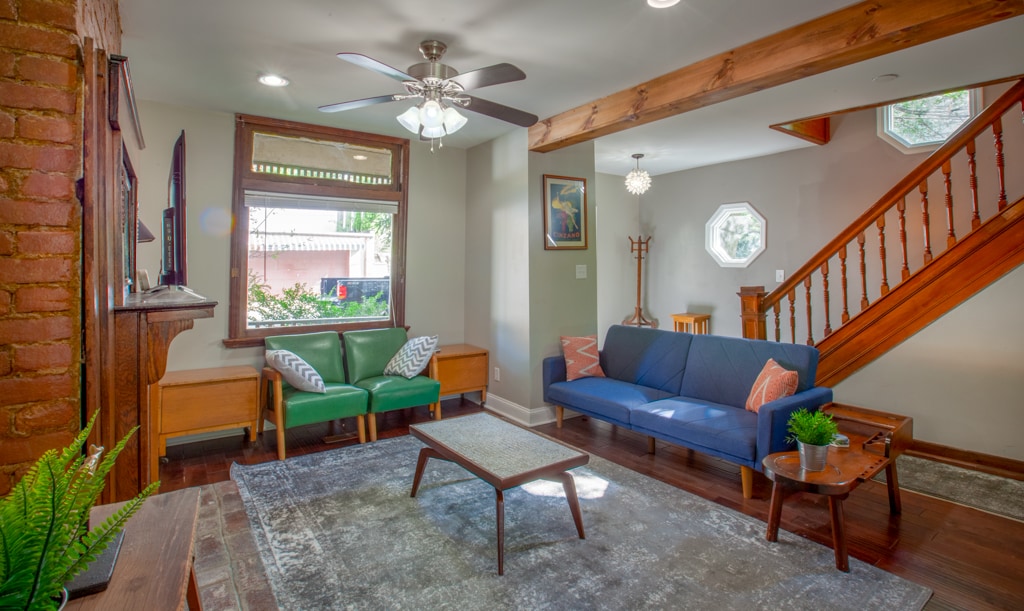 The living area features a blend of modern and rustic design, with a green armchair and a blue sofa positioned around a coffee table. Natural light enters through a window and a unique octagonal window, while warm wood tones are highlighted by wooden beams and a staircase in the background.