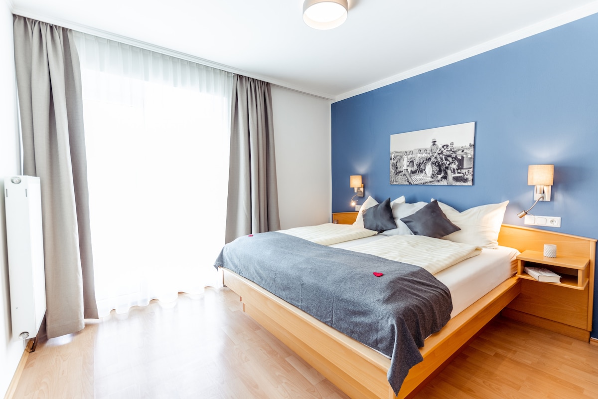 A spacious bedroom features a large bed with decorative pillows and a soft blanket. Natural light fills the room through expansive glass doors, complemented by light-colored curtains. The walls are adorned in a soothing blue hue, and nightstands with lamps are situated on either side of the bed.