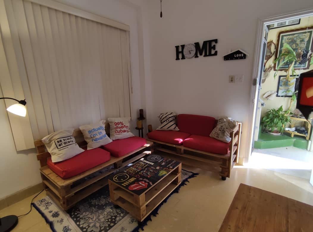 A cozy living area features a pallet-style seating arrangement with vibrant red cushions. Natural light flows in through a nearby door, illuminating the space. Decorative cushions and a small lamp add character to the room, which is adorned with wall art and personal touches.