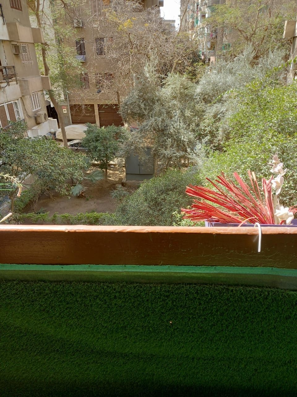 A balcony view showcases a serene green garden filled with trees and shrubs. The sight includes a patch of well-maintained grass, while colorful plants are positioned along the balcony railing, contributing to the tranquil atmosphere of the surroundings.