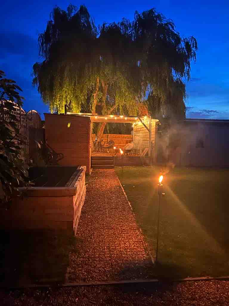 The outdoor area is softly illuminated by warm lights, showcasing a serene garden path leading to a cozy seating area under a shelter. A tall tree provides shade, and torchlights add ambiance to the inviting atmosphere of the space.