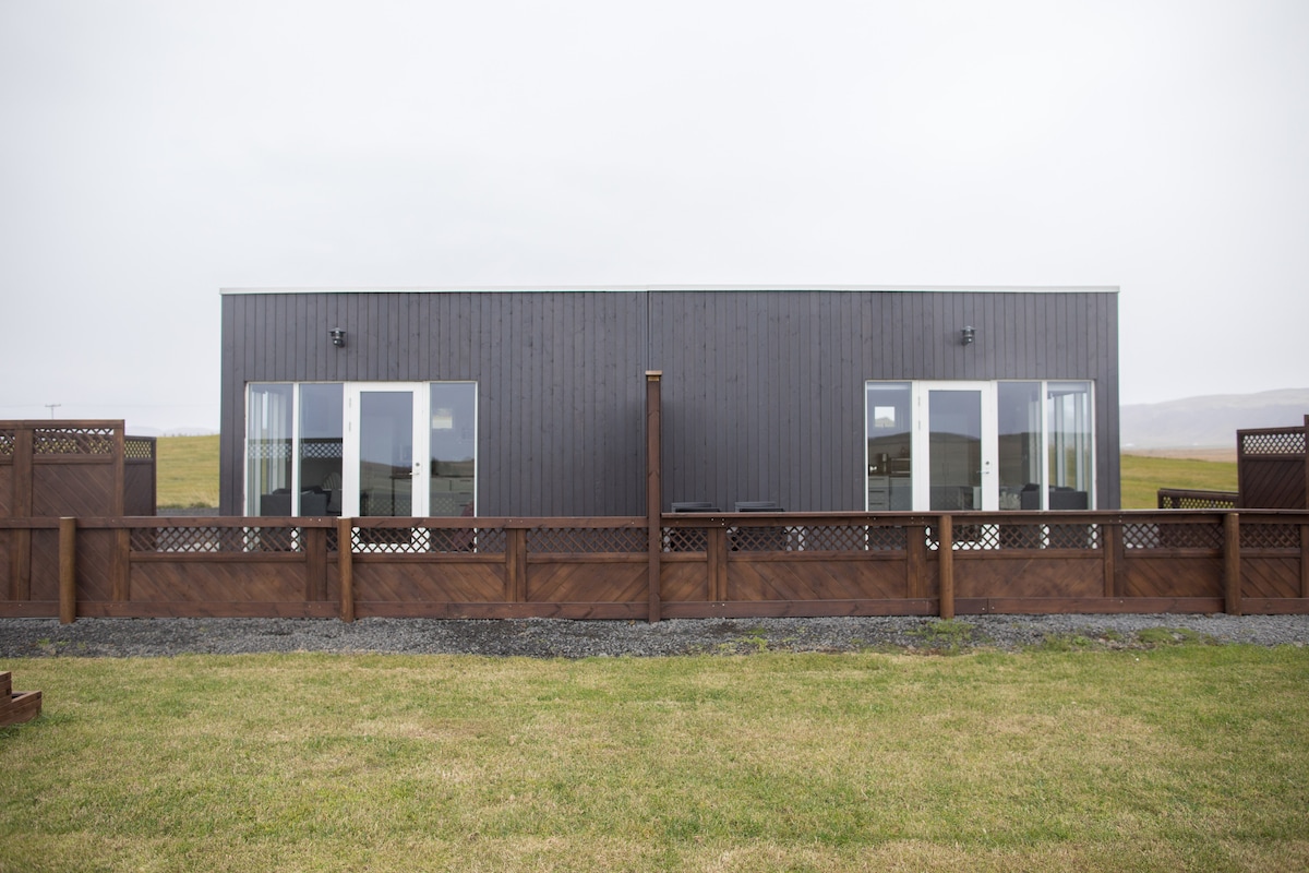 A modern, single-story building is depicted with a dark wooden exterior. Large glass doors provide access to the interior, surrounded by an enclosed yard featuring a wooden fence. Lush green grass extends in front, contributing to the serene landscape.