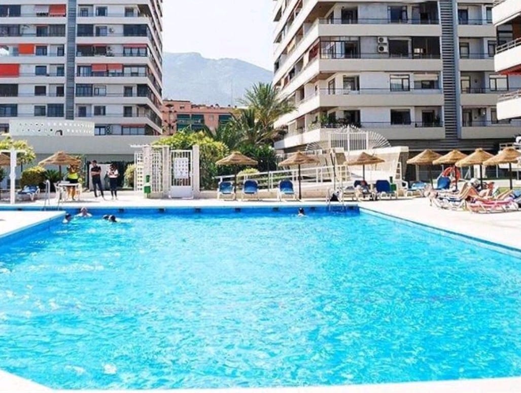 The image features a large outdoor swimming pool, surrounded by sun loungers and parasols. Several palm trees and landscaped gardens are visible in the background, alongside modern residential buildings that frame the area. Guests can be seen enjoying the pool on a sunny day.