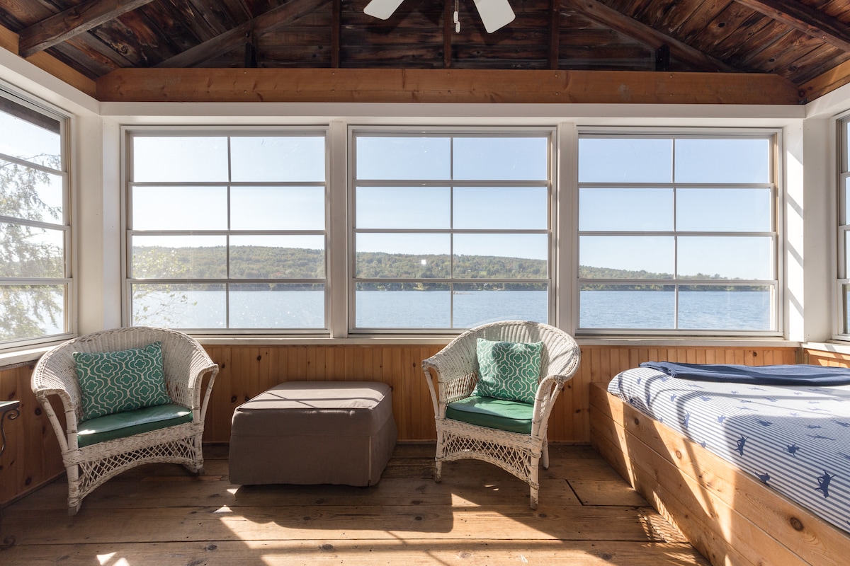 A screened porch showcases two wicker chairs with green cushions and a small ottoman, providing a comfortable seating area. Large windows frame expansive views of the serene lake, allowing natural light to fill the space. Wooden walls and flooring add warmth to the inviting environment.
