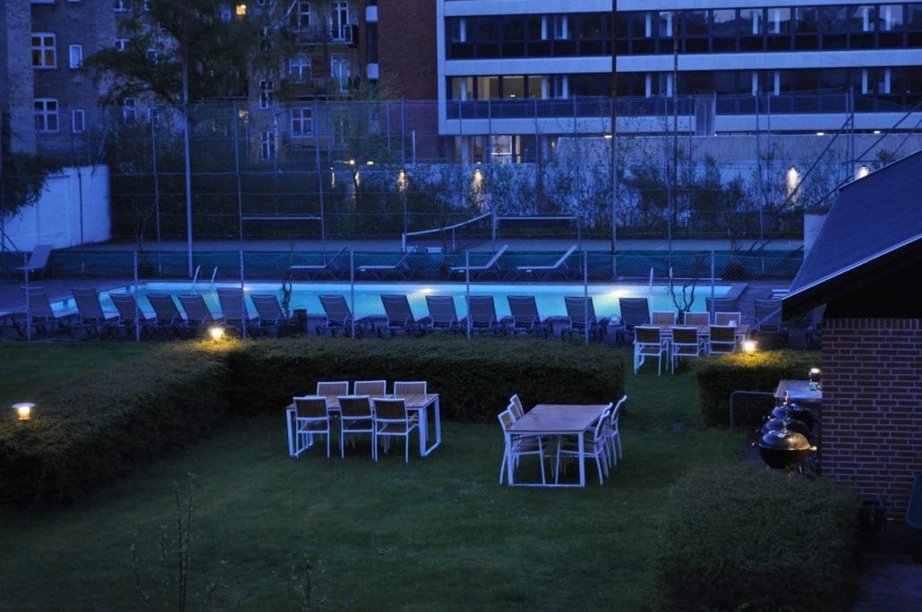An outdoor area is shown at night, featuring several illuminated tables and chairs arranged on a grassy lawn. A pool is visible in the background, surrounded by lounge chairs. Soft lighting enhances the setting, revealing nearby tennis courts.
