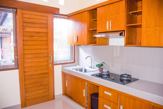 A modern kitchen features wooden cabinetry and a white countertop. A double sink and a gas stove are visible. Natural light enters through the window with horizontal blinds, creating a bright and functional cooking space.