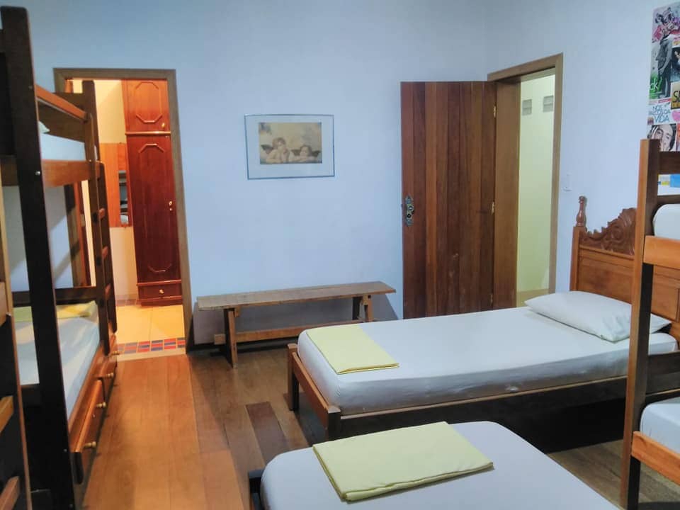 An inviting dormitory space accommodates multiple guests with three bunk beds and two single beds. A bench is positioned against the wall. Natural light enters through a nearby door leading to a bathroom, adding to the room's spacious feel.