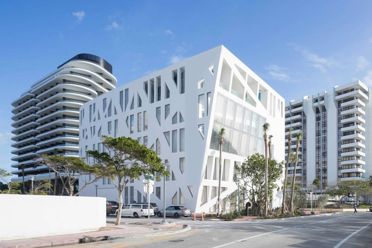 The contemporary architecture of the building features angular white facades with large windows, complemented by surrounding greenery. Nearby, sleek, modern structures reflect the stylish ambiance of Miami Beach. The image captures a bright blue sky, enhancing the striking design of the buildings.
