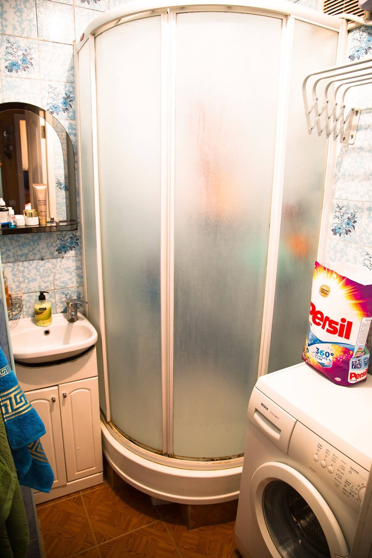 The bathroom features a compact shower cubicle with frosted glass doors. A small sink is positioned beside the shower, and a washing machine is located near the wall. Decorative tiles adorn the walls, complemented by a mirror above the sink.