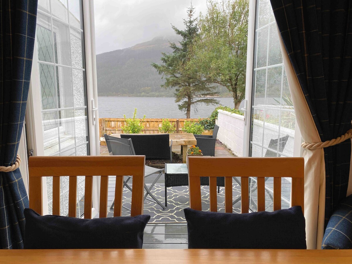 A view from the interior features French doors leading to a deck area. Outdoor seating is positioned facing Loch Long and the surrounding landscape, framed by trees. Natural light is filtered through curtains with a tartan pattern, enhancing the inviting space.