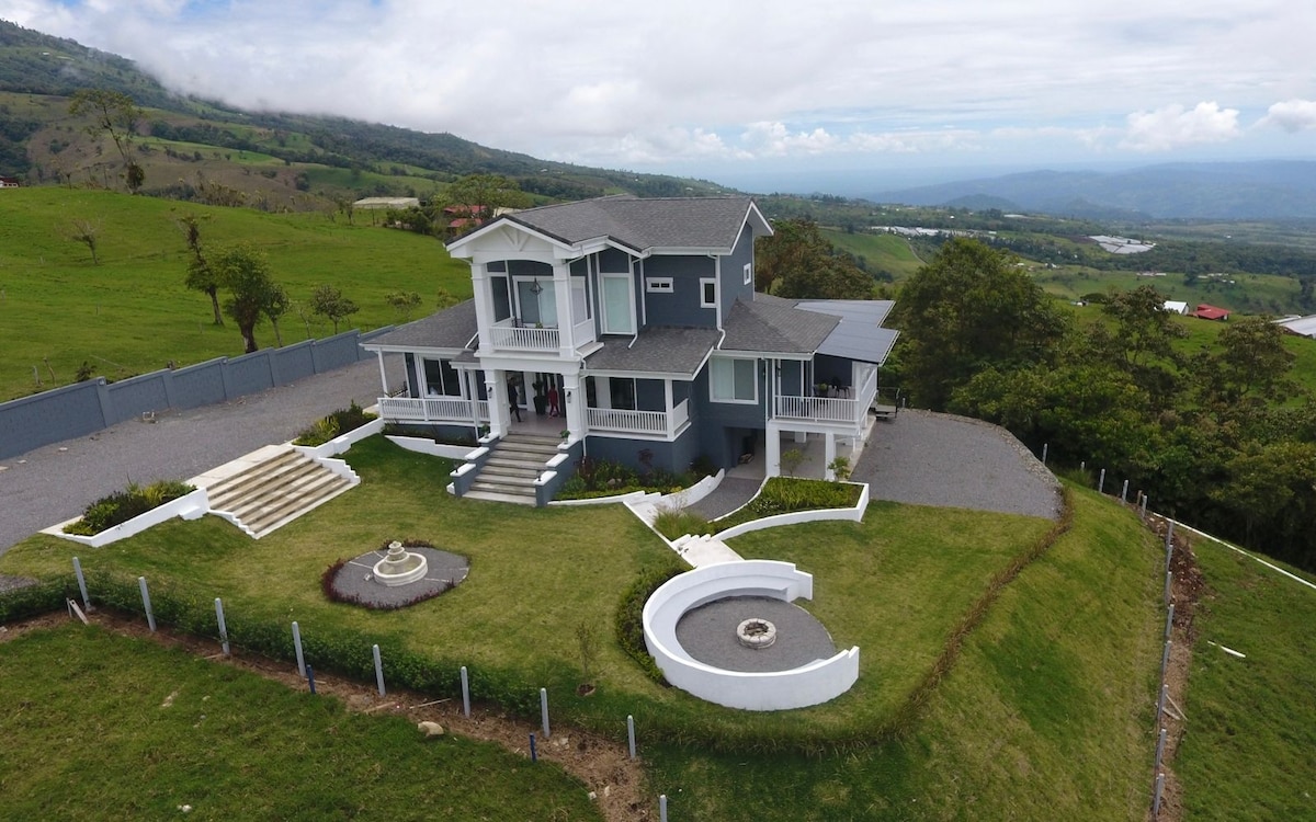 A grand two-story house is surrounded by lush green hills, with neatly landscaped gardens and a circular driveway. Expansive windows reflect the natural landscape, and pathways lead to a central decorative feature in the yard, inviting a sense of tranquility.
