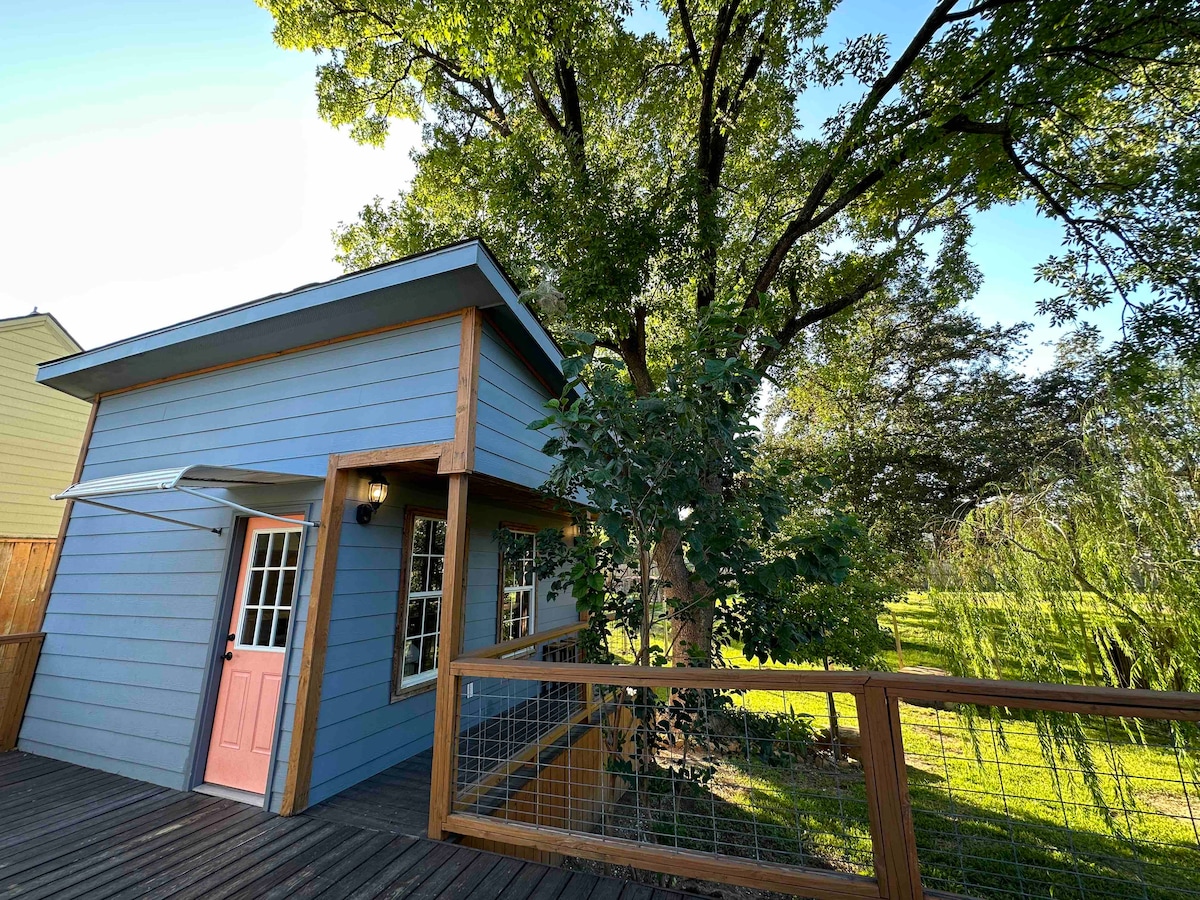 The exterior of the guest house is highlighted, showcasing a light blue façade with a welcoming pink door. Lush green trees and plants surround the structure, providing a sense of tranquility. A wooden deck with railing leads up to the entrance.