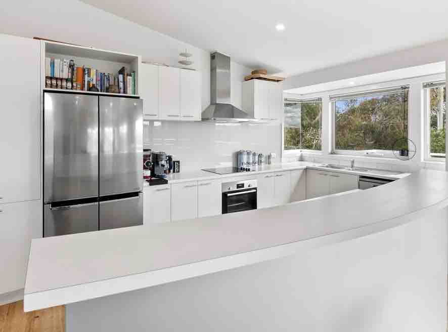 A modern kitchen is presented with stainless steel appliances, including a refrigerator and oven. A white countertop offers ample space for meal preparation. Natural light fills the room through multiple windows, illuminating the sleek cabinetry and a well-organized shelf with books and kitchen items.