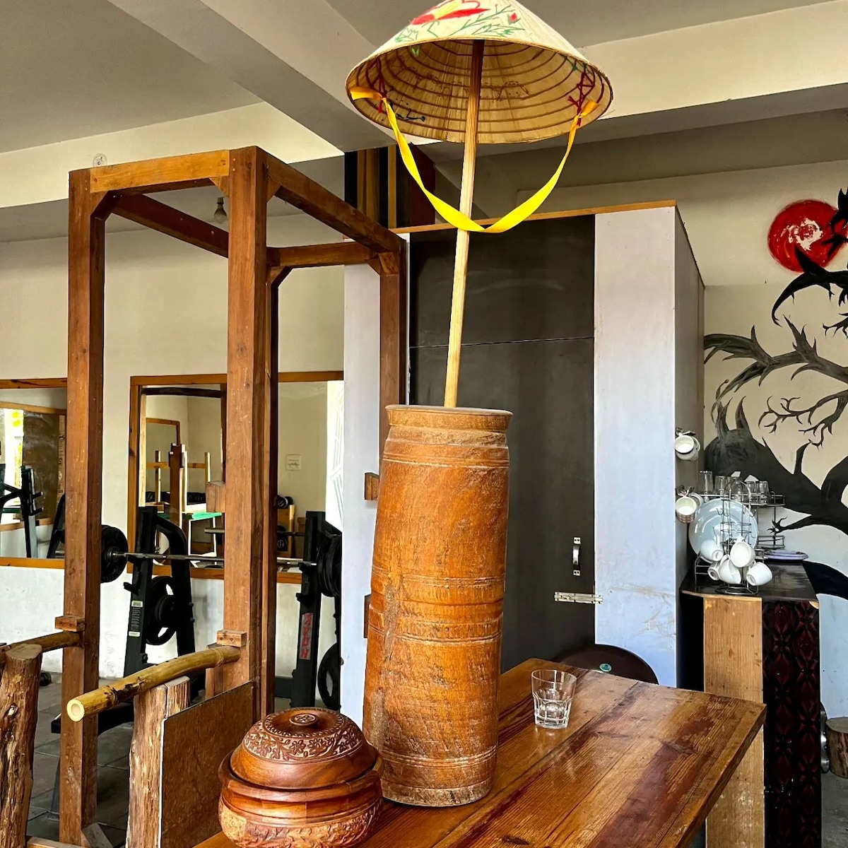 A wooden table displays a traditional hat mounted on a tall wooden vase. Textured elements from the furniture create a rustic ambiance. In the background, gym equipment is partially visible, complemented by decorative wall artwork, showcasing a blend of functionality and style.