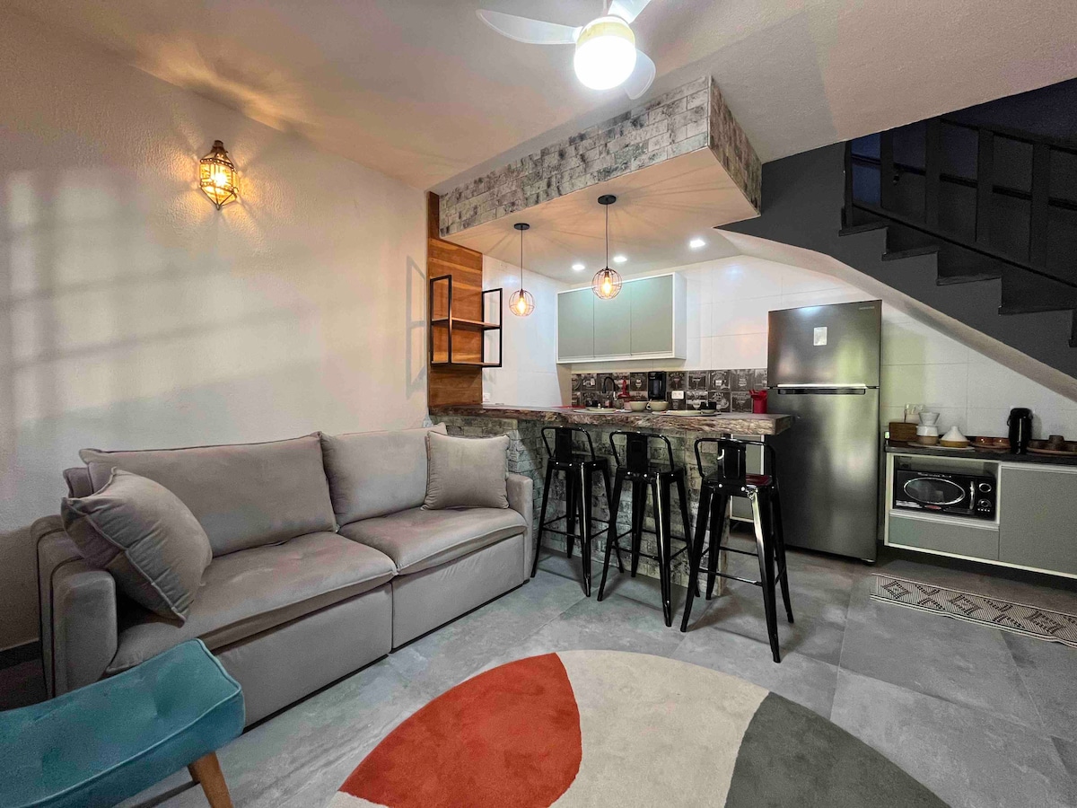 A cozy living space is highlighted with a light gray sofa and a circular rug featuring earthy tones. A kitchen bar with high stools is positioned against a wall of modern appliances, including a refrigerator, along with bright pendant lighting providing a warm ambiance.