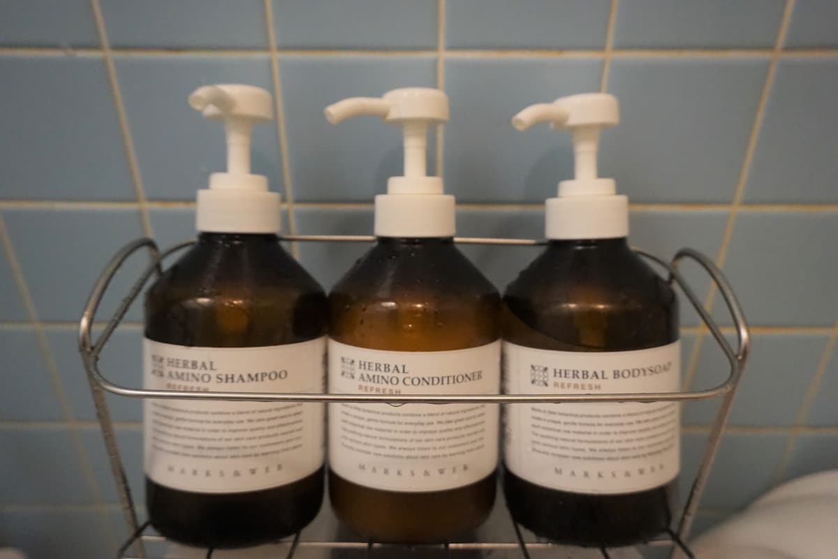 Three brown glass bottles with pump dispensers are arranged on a metal rack. Each bottle is labeled with product information, including herbal shampoo and body soap, set against a light blue tiled wall.