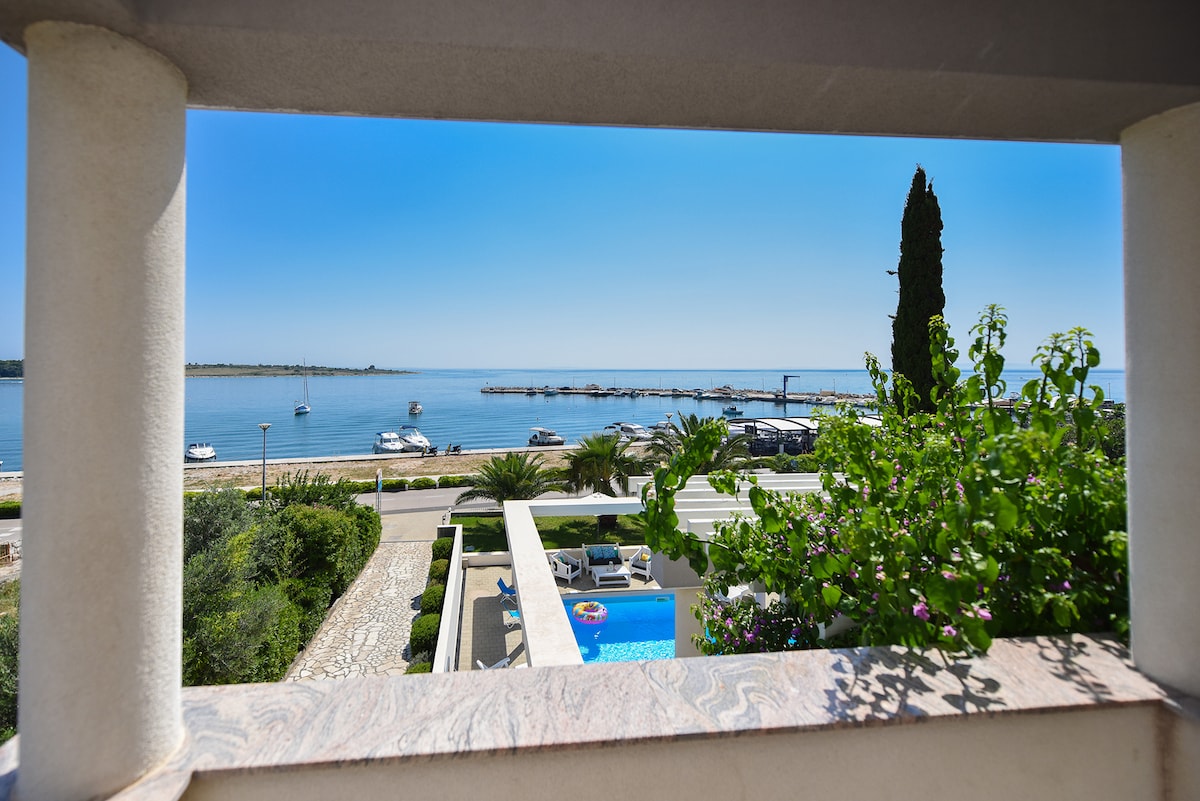 A tranquil coastal view is framed by a balcony. The scene displays serene waters with boats anchored in the distance, complemented by lush greenery and palm trees lining the waterfront. Sunlight brightens the landscape, enhancing the calm ambiance.