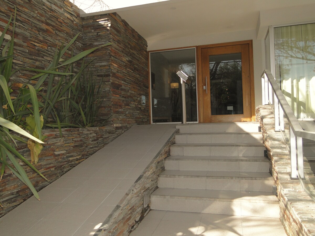 An inviting entrance features a ramp alongside stone walls adorned with greenery. Smooth, wide steps lead to a set of wooden doors that facilitate entry into the home. Natural light enhances the clarity of the space.