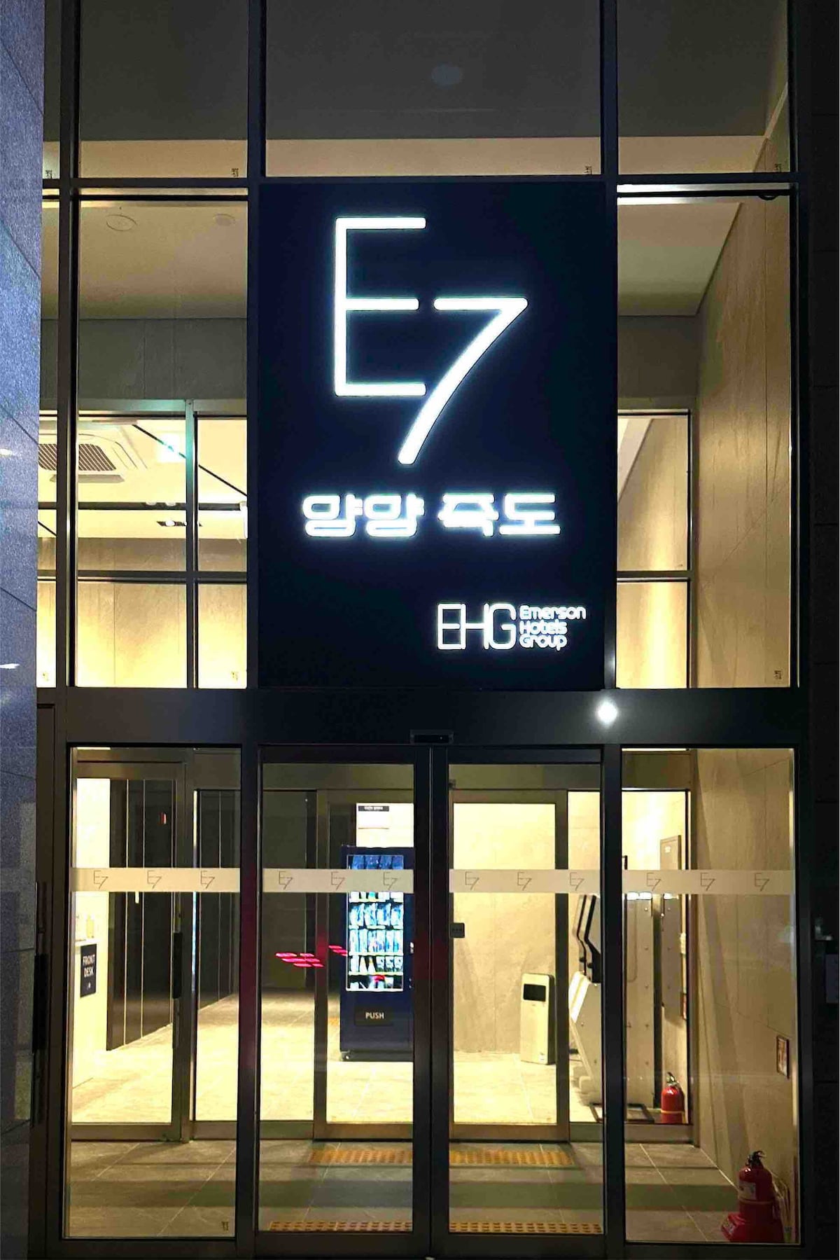 The entrance to the building is showcased, featuring large glass doors with a modern design. A bright illuminated sign displays the name 'E7' prominently above. The surroundings are minimalistic, with soft lighting enhancing the welcoming atmosphere.