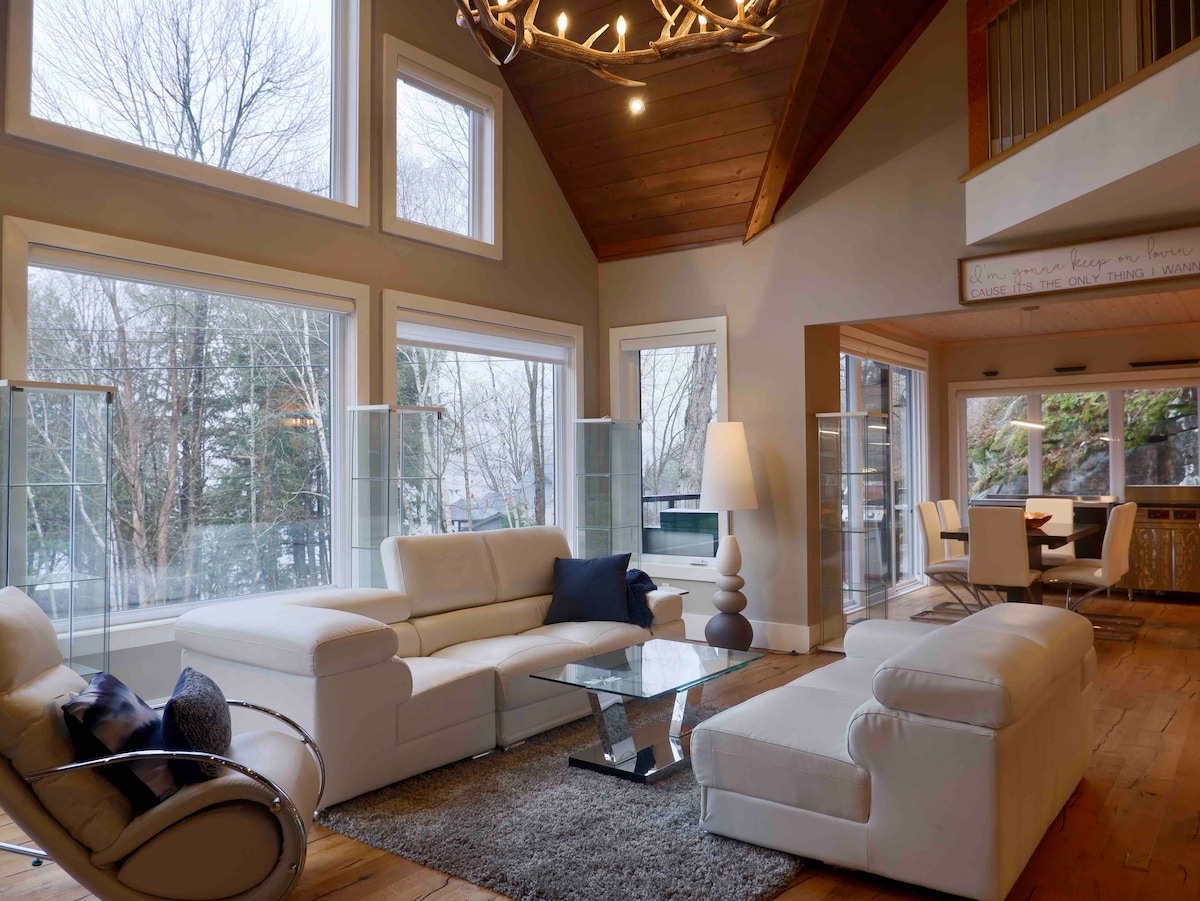 A spacious living area is highlighted by large windows that allow natural light to fill the room. Comfortable white sofas are arranged around a glass coffee table, with a cozy area rug beneath. The high wooden ceiling adds a sense of openness, complemented by modern decor.