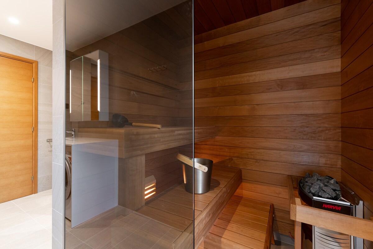 A soothing sauna is displayed with wooden benches and soft ambient lighting. A bucket and ladle are positioned on one side, while a heater filled with stones sits in the corner, providing warmth and relaxation.