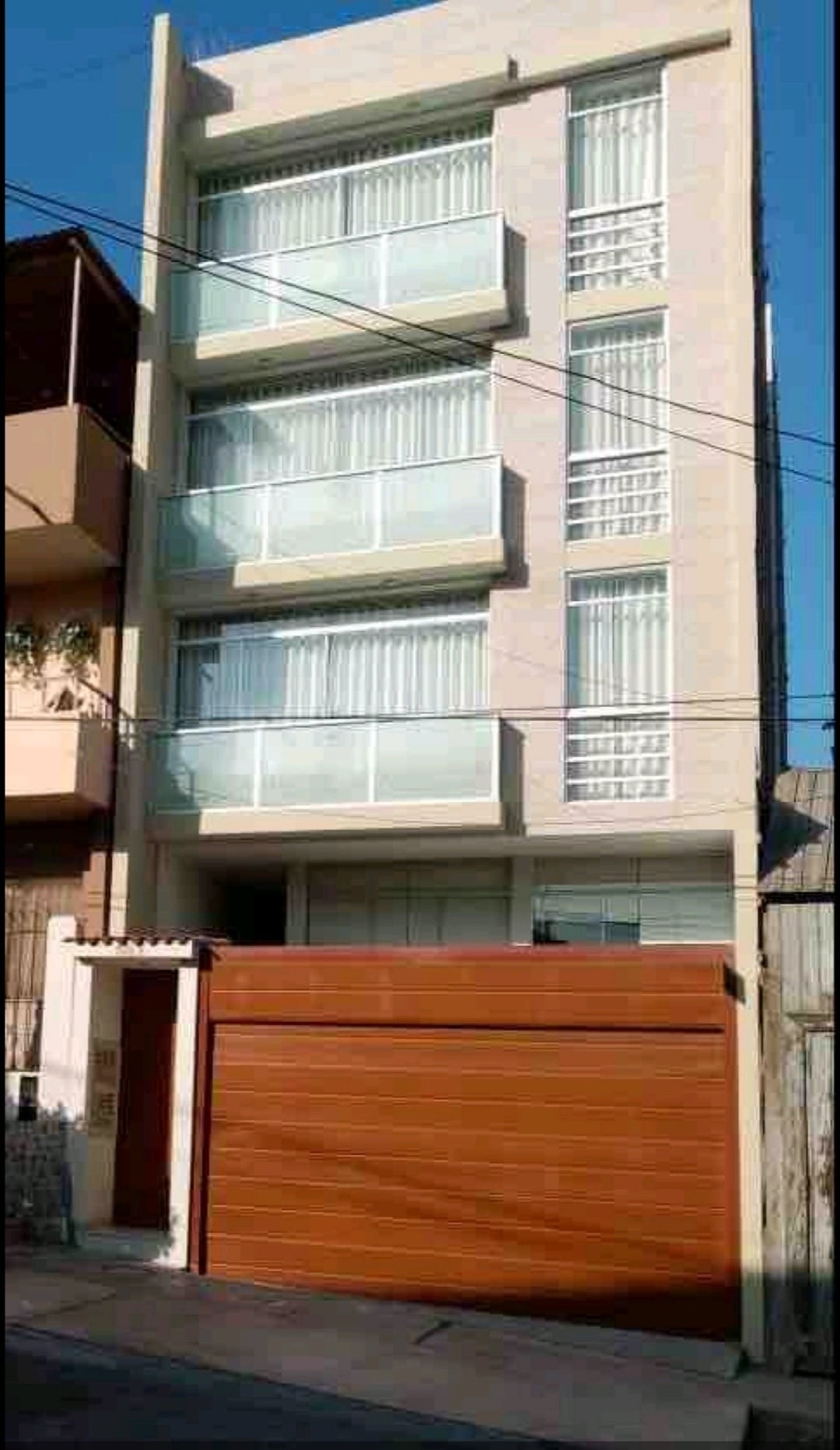 A modern multi-story building features a facade with large glass windows and balconies. The entrance is accompanied by a wooden garage door. Sunlight reflects off the smooth surfaces, highlighting the building's contemporary design.
