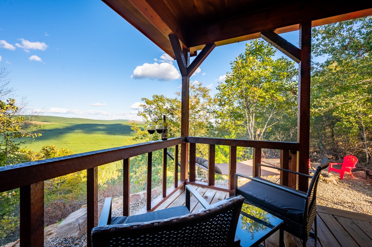A wooden deck is shown with two wicker chairs positioned for views of the surrounding trees and mountains. Natural sunlight brightens the scene, highlighting the expansive landscape and a vibrant red chair in the distance.