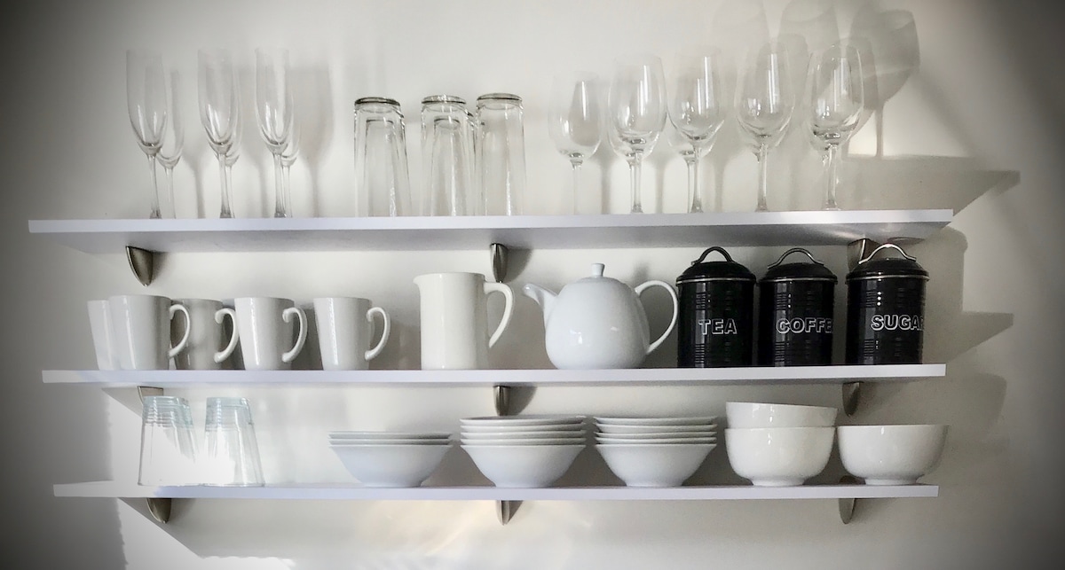 A wall-mounted shelf displays an organized arrangement of glassware and dishware. Glasses and mugs are positioned on the top shelf, while the bottom shelf features white bowls and cups. Three black canisters labeled 'TEA,' 'COFFEE,' and 'SUGAR' are also included.