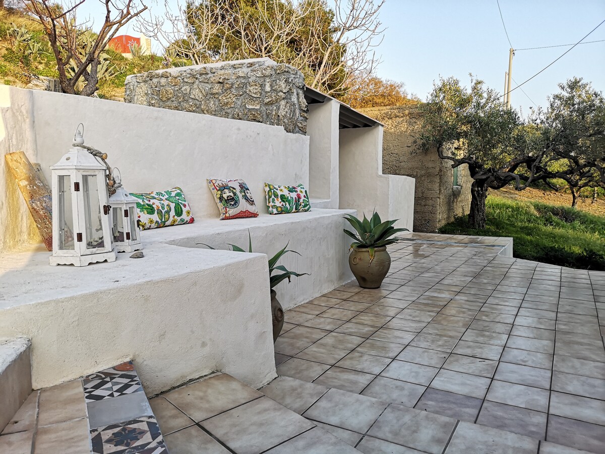 A sunlit outdoor patio area features tiled flooring and built-in seating against a stone wall. Colorful cushions add comfort, while decorative lanterns offer a cozy touch. Potted plants enhance the garden setting, with olive trees visible in the background, contributing to the serene ambiance.