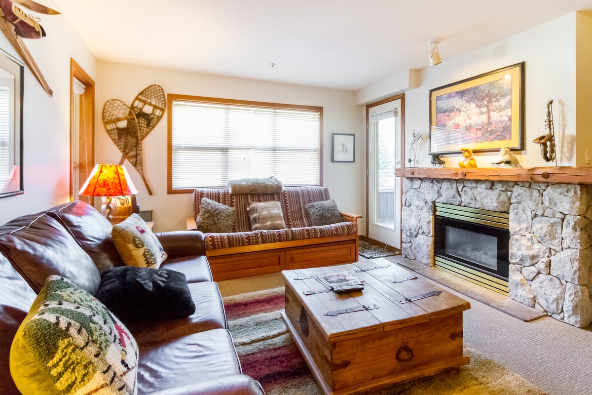 A comfortable living room is featured, showcasing a leather sofa and a wooden coffee table. Natural light filters in through the windows, illuminating the stone fireplace. A cozy seating area with a striped sofa and decorative snowshoes on the wall adds character to the space.